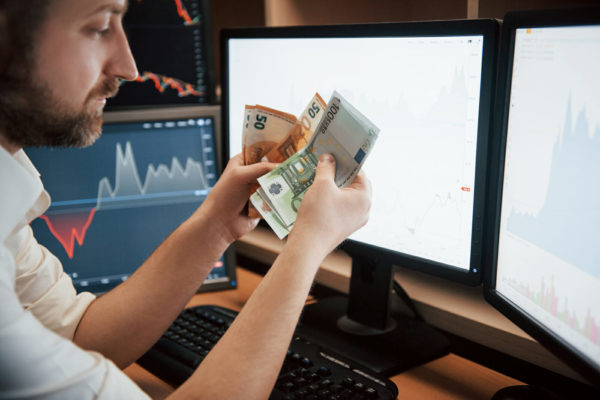 Time to get paid. Bearded man in white shirt holds earned money in the office with multiple computer screens in index charts.