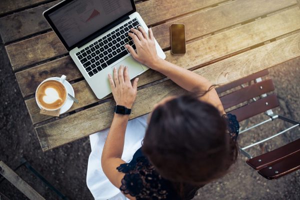 Top view of female using her laptop at a cafe. Overhead shot of young woman sitting at a table with a cup of coffee and mobile phone surfing the net on her laptop computer.