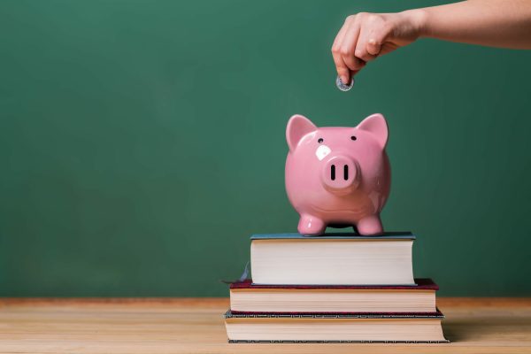 Person depositing money in a pink piggy bank on top of books with chalkboard in the background as concept image of the costs of education
** Note: Soft Focus at 100%, best at smaller sizes